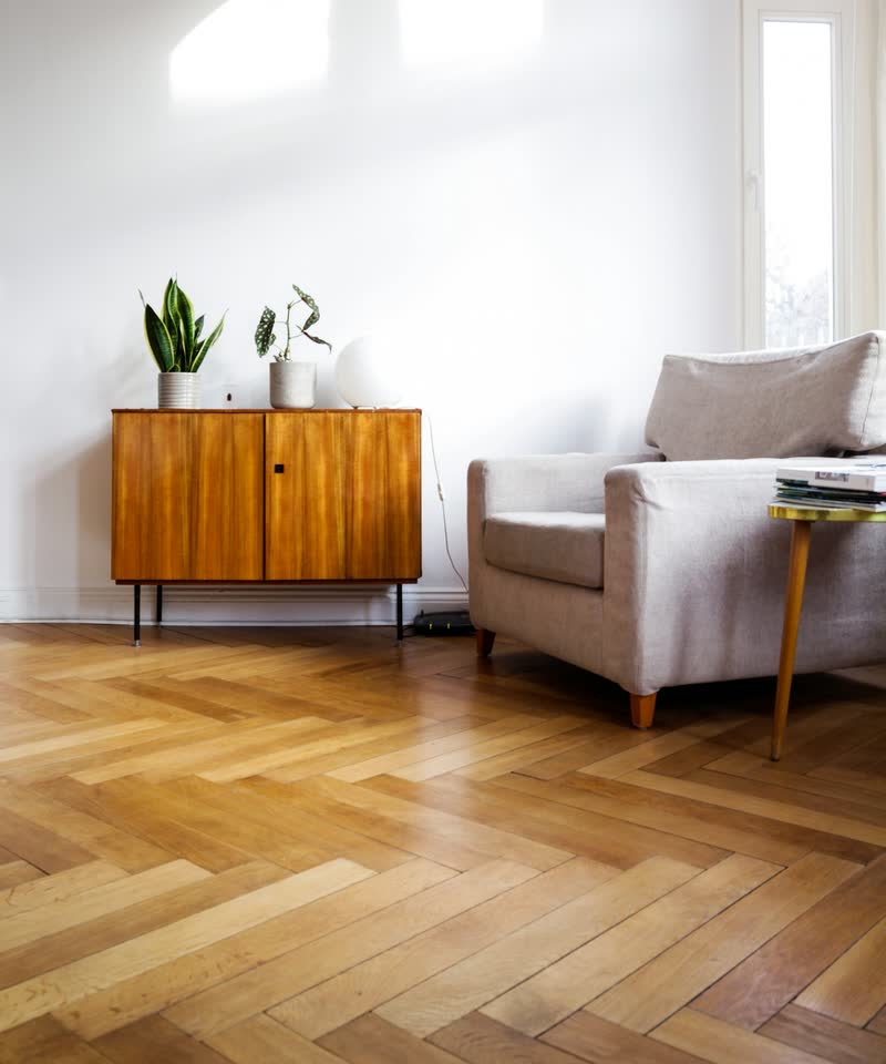 A sunny living room with a herringbone wood floor, a wooden sideboard and an armchair.