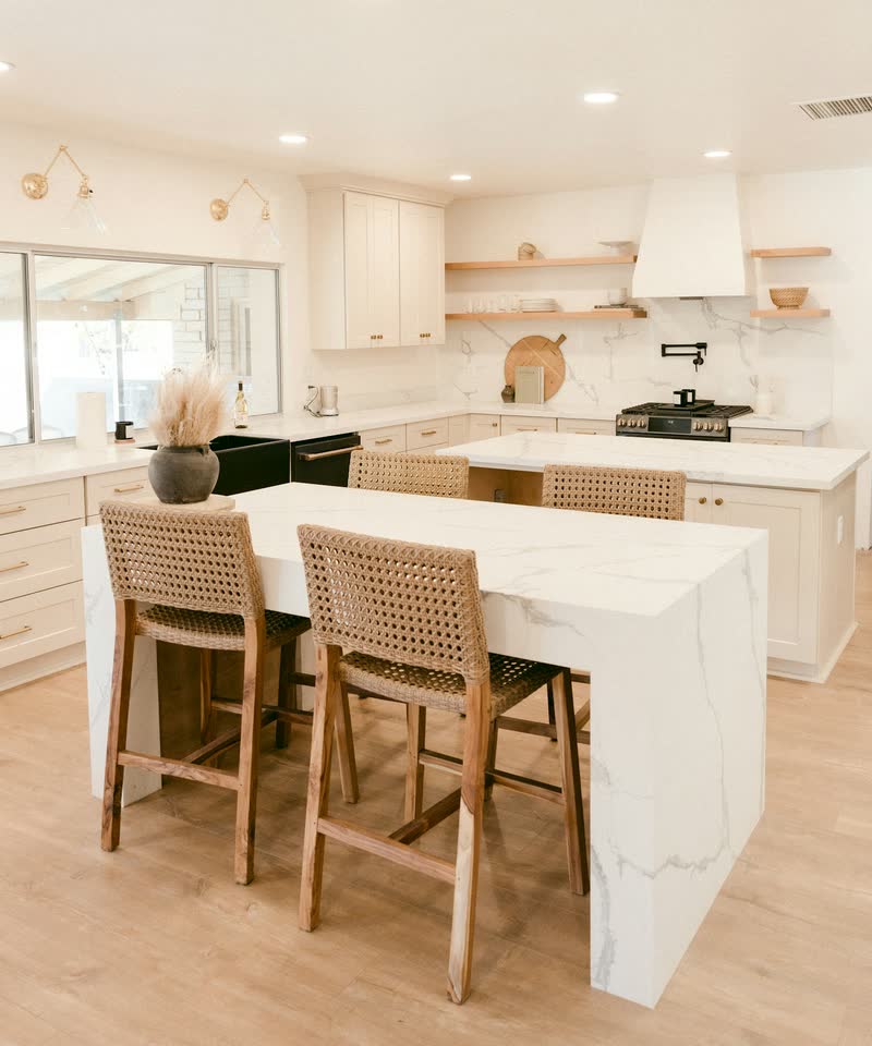 A white kitchen with a large pale stone island and woven bar stools.