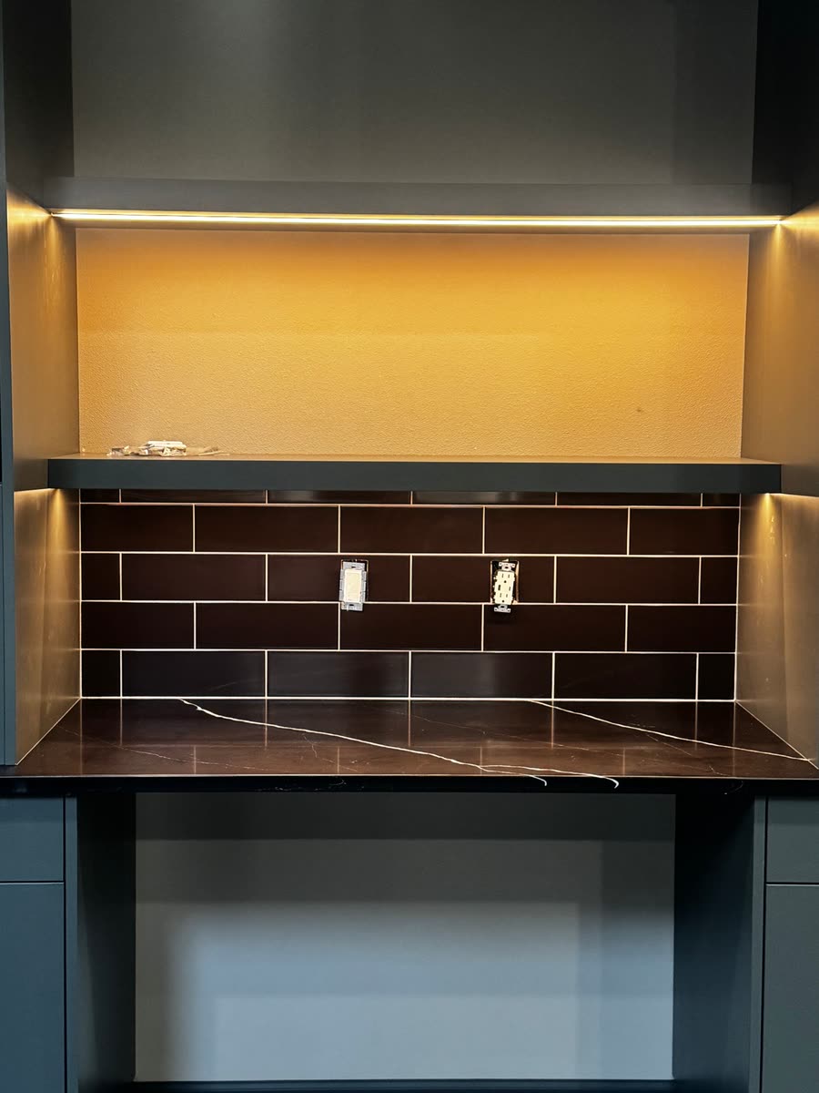 A bar counter in dark veined stone under a brown tile splash, with lighting above.