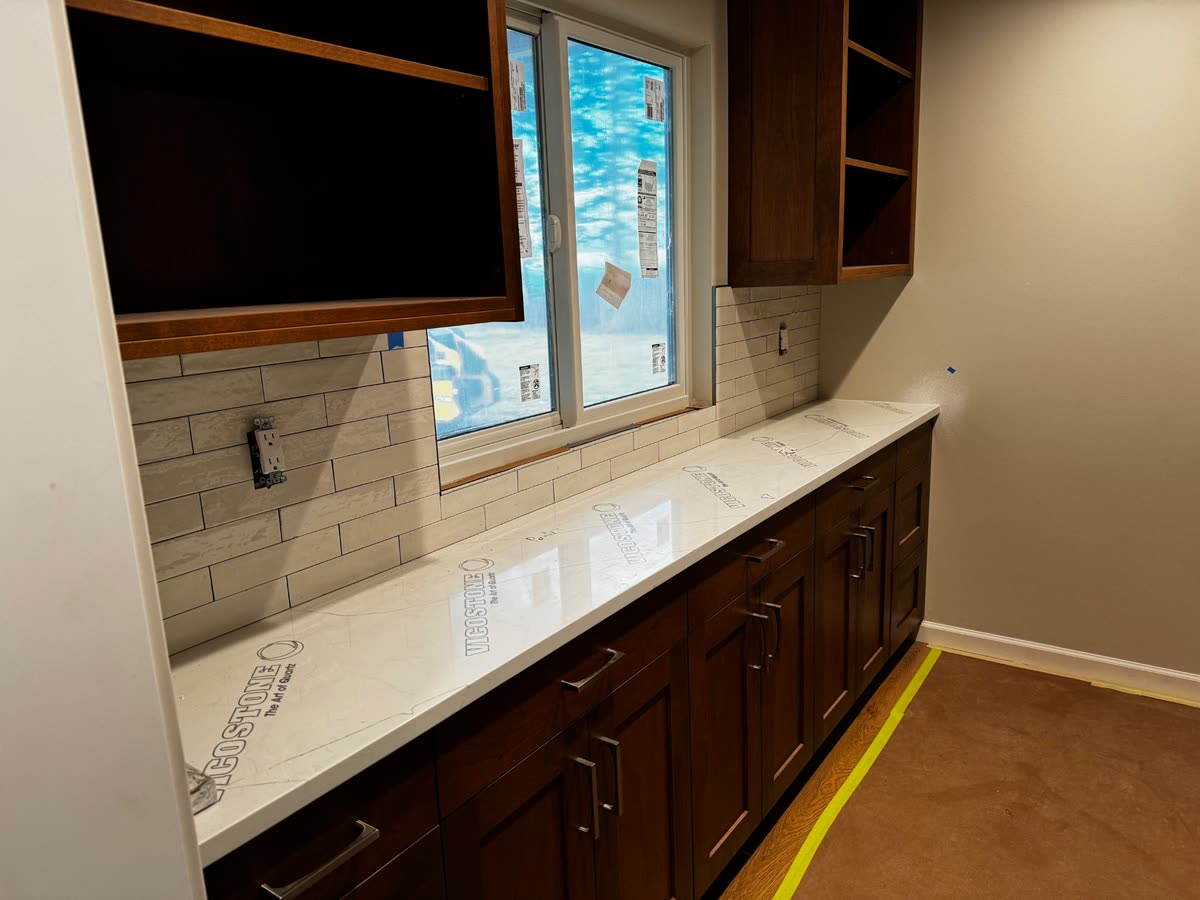 A long pantry counter in pale quartz with the maker's protective film still on it, subway tile behind.