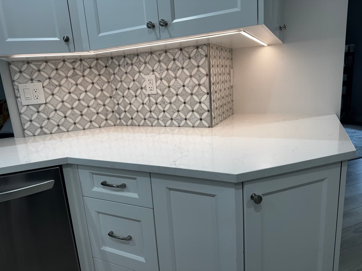 A circular-patterned marble mosaic splash above a pale stone counter, under-cabinet lighting on.