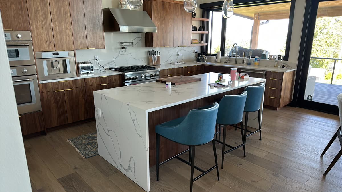 A kitchen with a wide-plank wood floor, dark wood cabinets and a pale stone island with a waterfall edge.