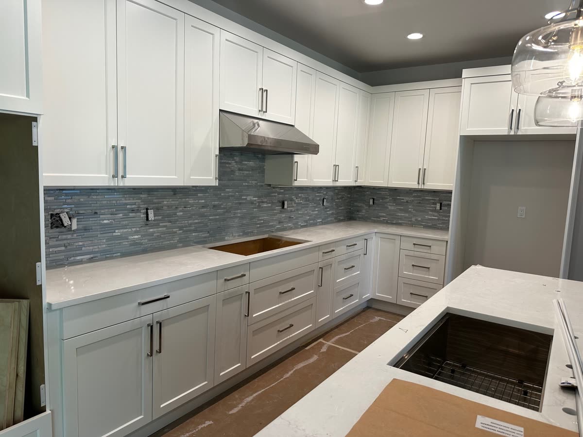 A white kitchen with a narrow glass mosaic splash and pale stone counters, sink cut out ready.